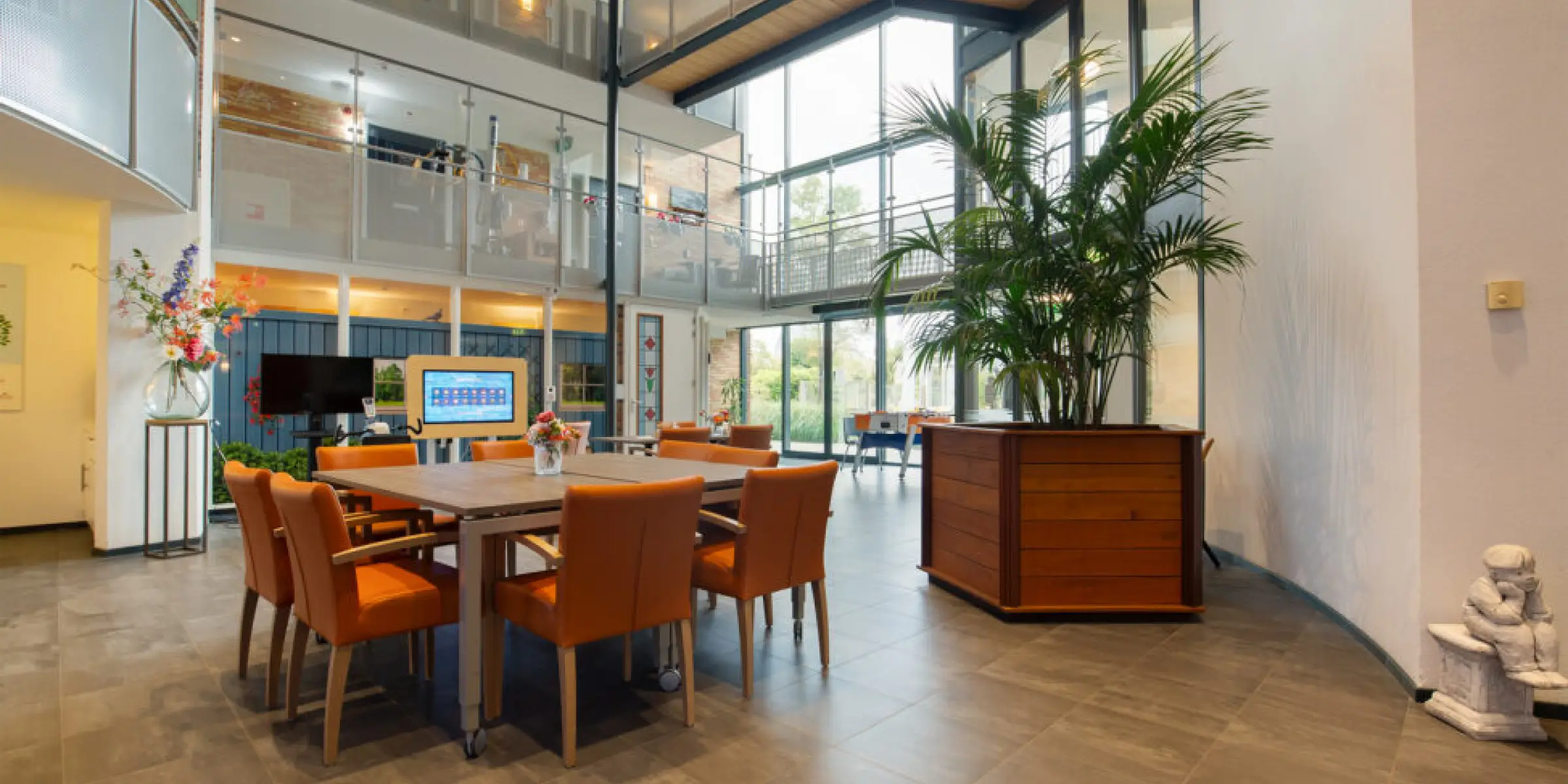 Geurbeleving-in-de-zorg-gezellige-gemeenschappelijke-ruimte | allsens b. V. Gemeenschappelijke ruimte in een zorginstelling met houten tafel, oranje stoelen, verse bloemen en een grote kamerplant; geurbeleving helpt hier om een huiselijke sfeer te creëren.