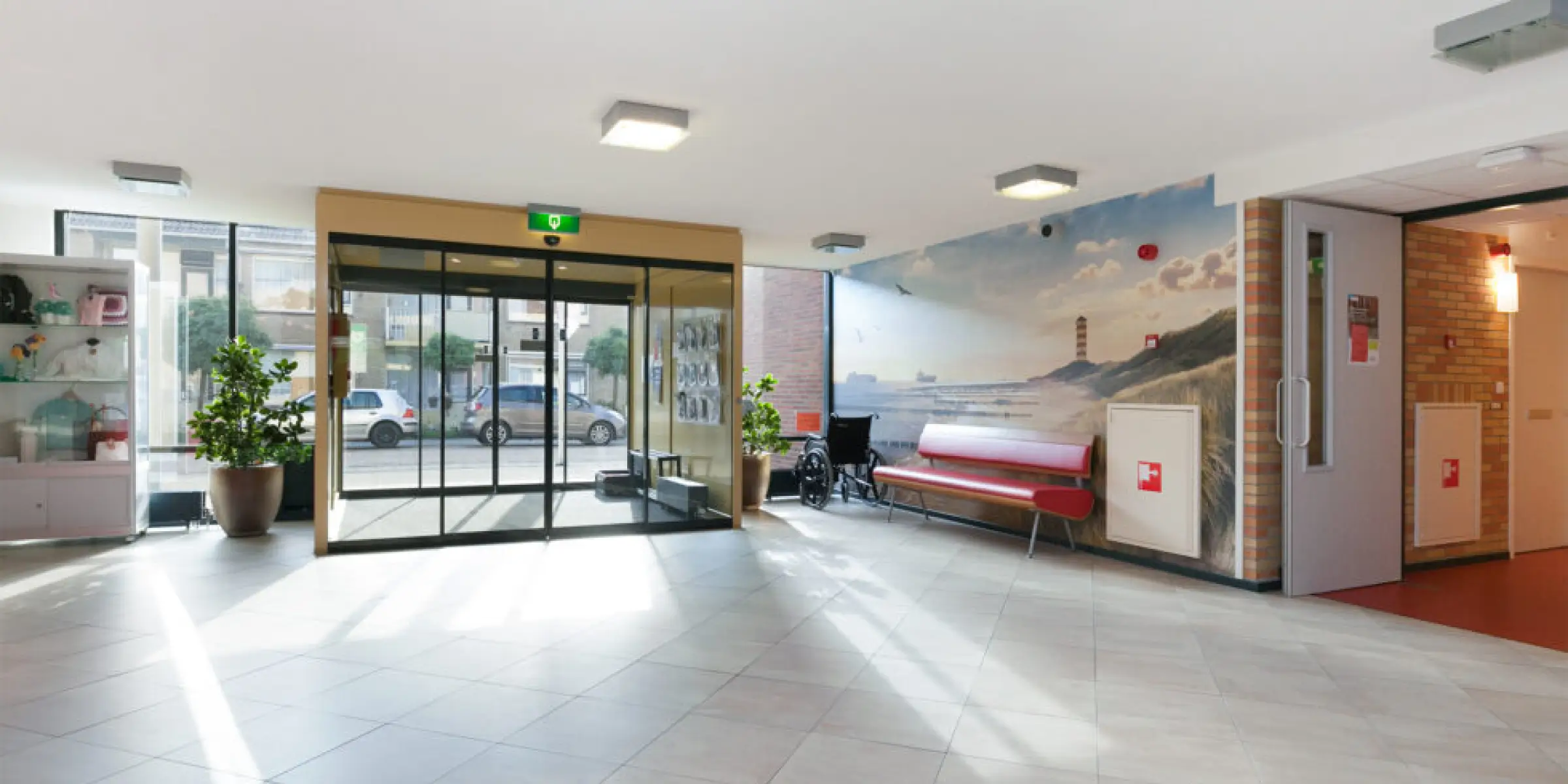 Geurbeleving-in-de-zorg-lichte-lobby-van-zorginstelling | allsens b. V. Lichte lobby van een zorginstelling met glazen schuifdeuren, een rode bank, een rolstoel en groene planten; hier kan geurbeleving in de zorg een warme en gastvrije entree creëren.