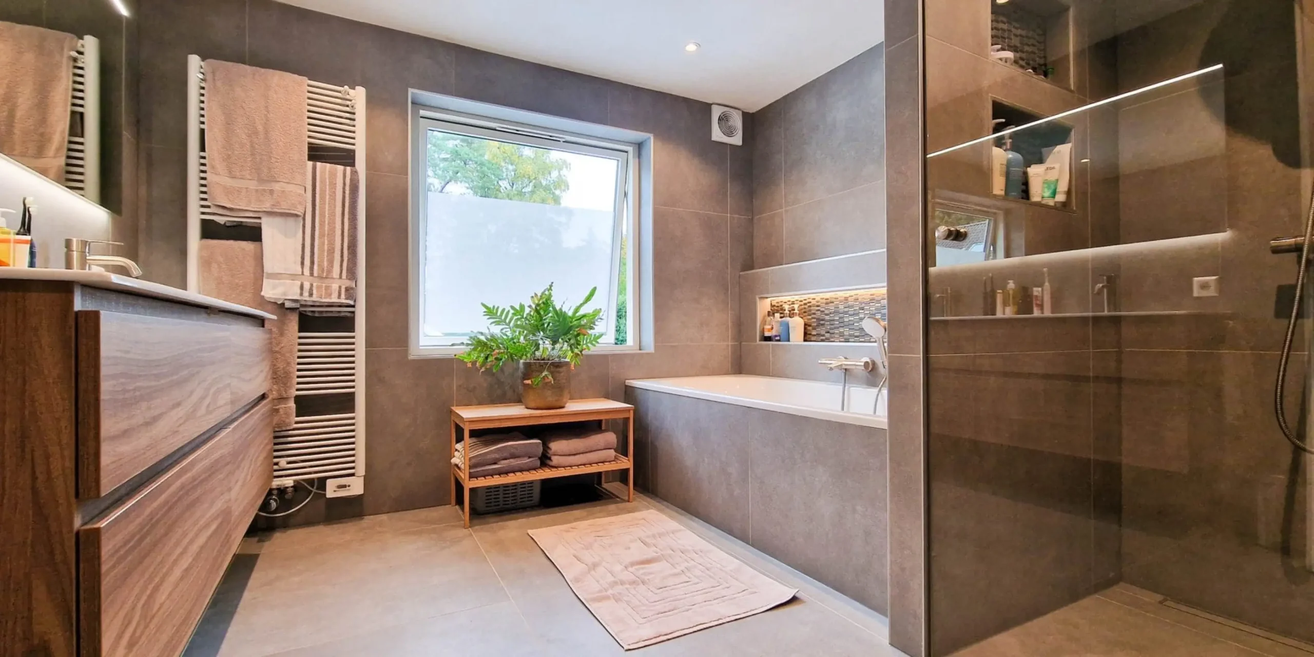 Geurbeleving-showroom-moderne-badkamer-met-inloop-douche | allsens b. V. Stijlvolle badkamer met houten wastafelmeubel, inloopdouche met glazen wand, ligbad en een groene plant, een warme ruimte die geuren tot hun recht laat komen.