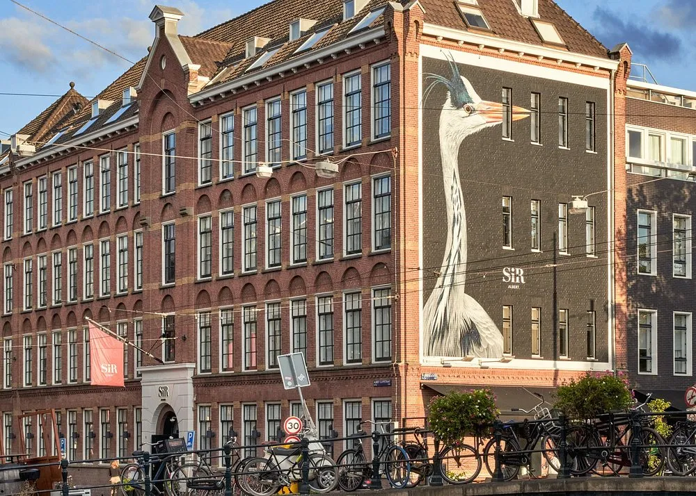 Bakstenen hotelpand met grote ramen, muurschildering van een ooievaar en sir‑hotels‑vlaggen; geuren dragen bij aan een uitnodigende entree.