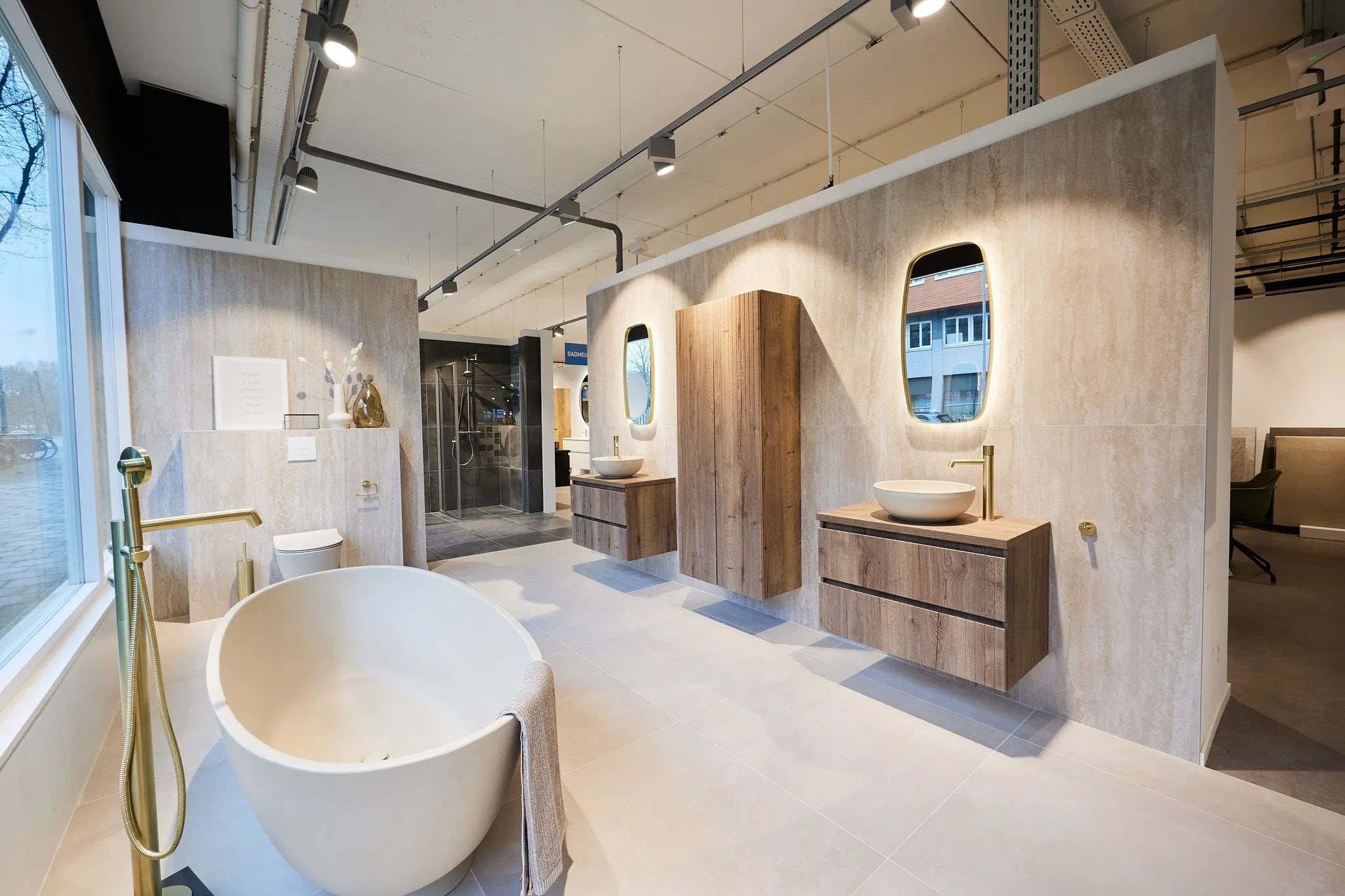 Geurbeleving-verrijkt-de-showroomervaring-bij-sanidirect-luxe-badkamer-showroom | allsens b. V. Luxe badkamershowroom met vrijstaand wit bad bij een raam, houten wastafel en spiegel; geurbeleving creëert een uitnodigende sfeer voor klanten.