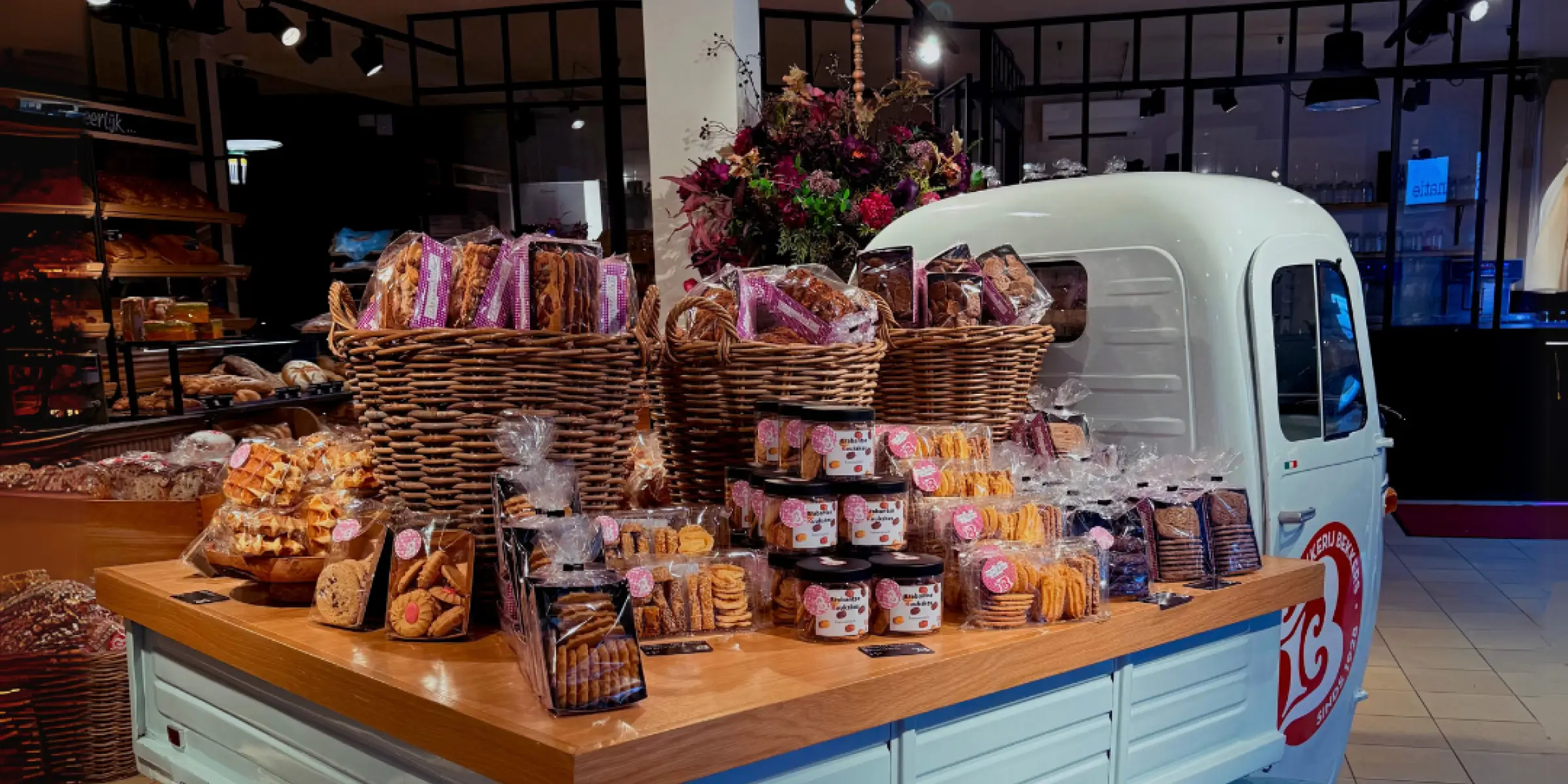 Binnenpresentatie met een vintage bestelbus vol rieten manden brood en gebak in een sfeervolle bakkerij; de geur van vers brood vult de ruimte.