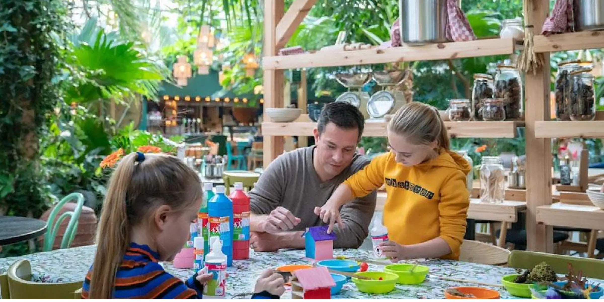 Vader helpt twee jonge meisjes met knutselen aan een tafel vol papieren en potloden in een groene indoor‑omgeving; de geurbeleving maakt de sfeer huiselijk.