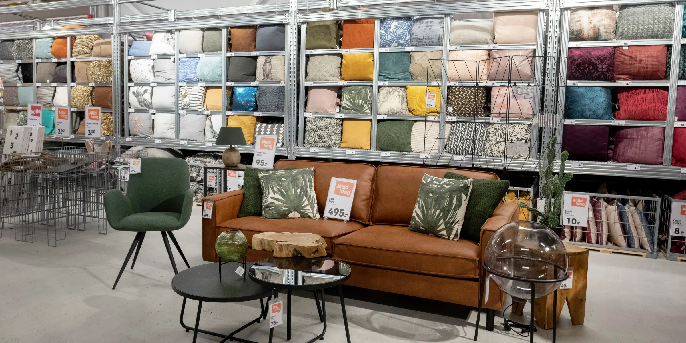 Geurmarketing-bij-woonwinkel-kwantum-versterkt-beleving-bank-groene-stoel | allsens b. V. Gezellige presentatiehoek in de kwantum‑woonwinkel met een cognackleurige bank, groene stoel en wand vol kussens; geurmarketing versterkt hier de winkelbeleving.